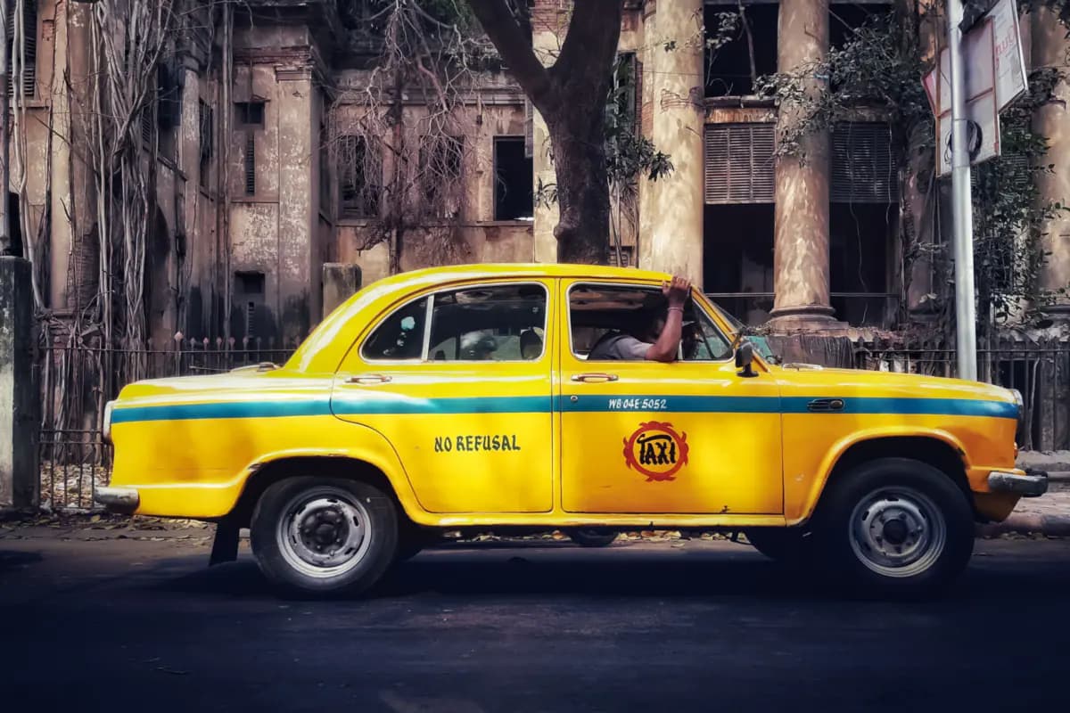 A yellow taxi with a blue stripe in front of an old colonnaded building overgrown with hanging roots on a Kolkata street