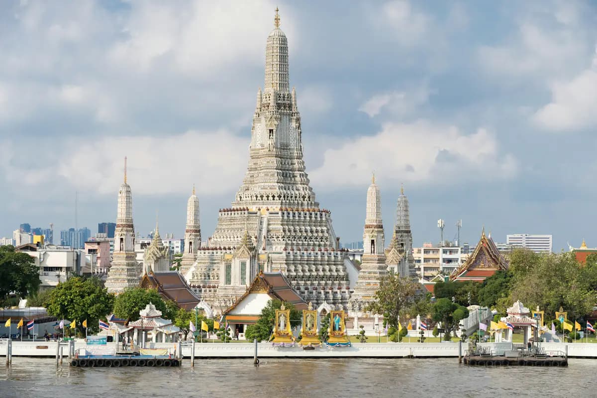 Wat Arun's central prang and smaller towers behind a riverside wall with flags, seen across the Chao Phraya in Bangkok