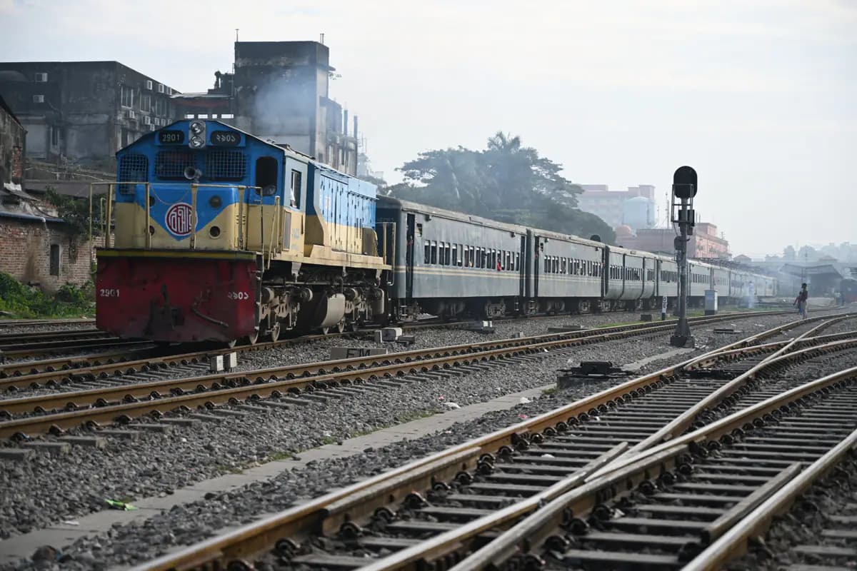 A blue and yellow diesel locomotive pulling passenger coaches past the tracks at Chattogram station
