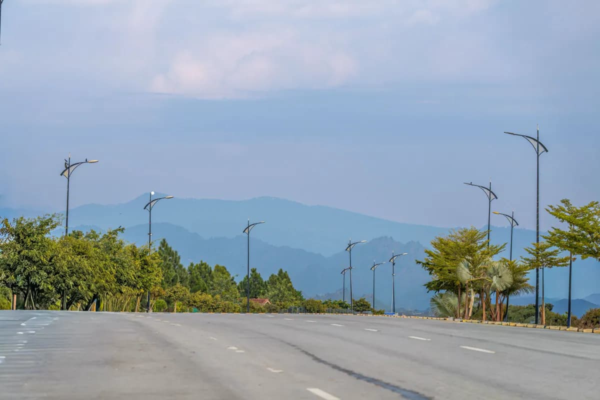 A wide, empty avenue with tall street lamps and trees leading towards hazy blue hills in Islamabad