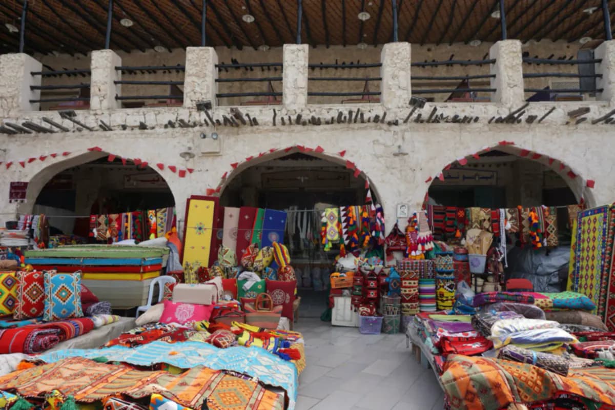 Woven cushions, rugs and bags piled on stalls under the arches of a market building in Souq Waqif
